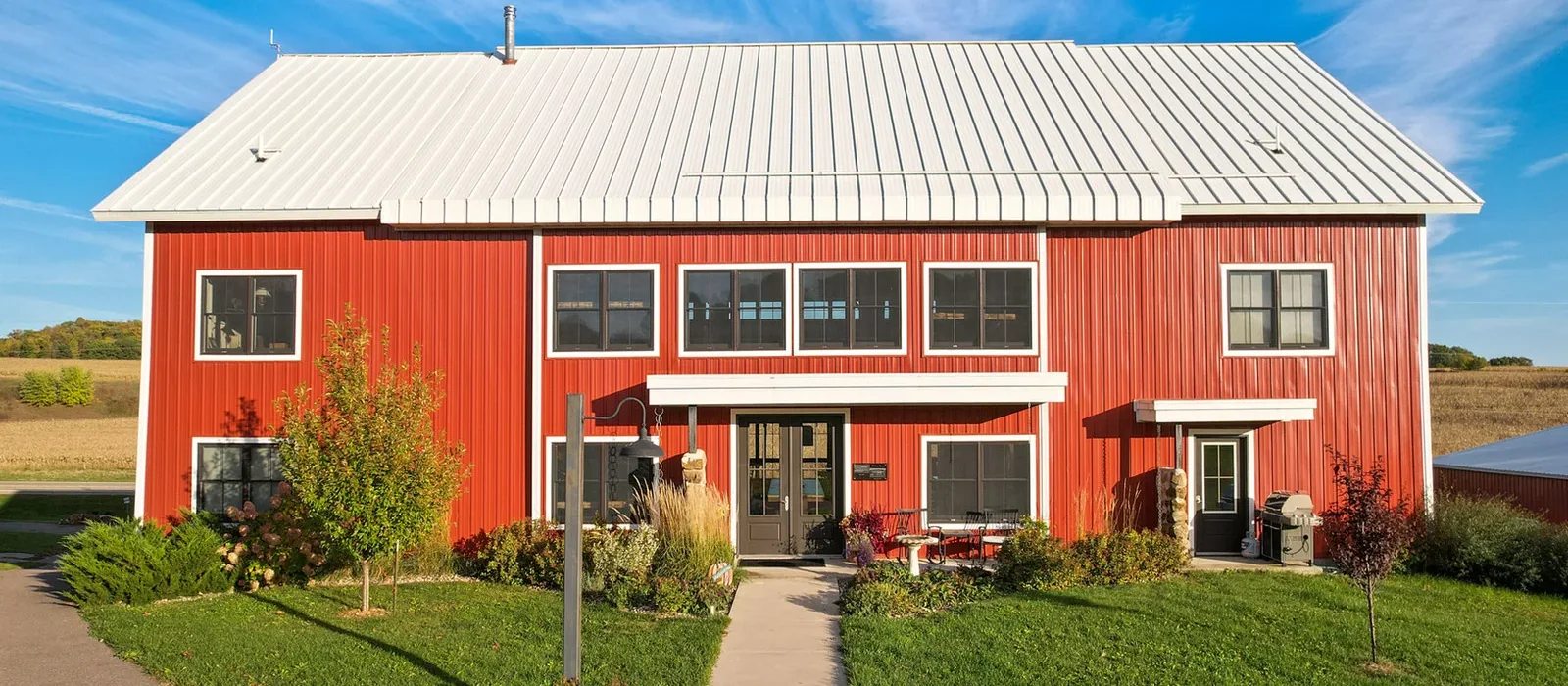 The renovated Pelton barn-home shown head-on under a clear sky — full red facade, white metal roof, both stories of windows, and the lawn in front